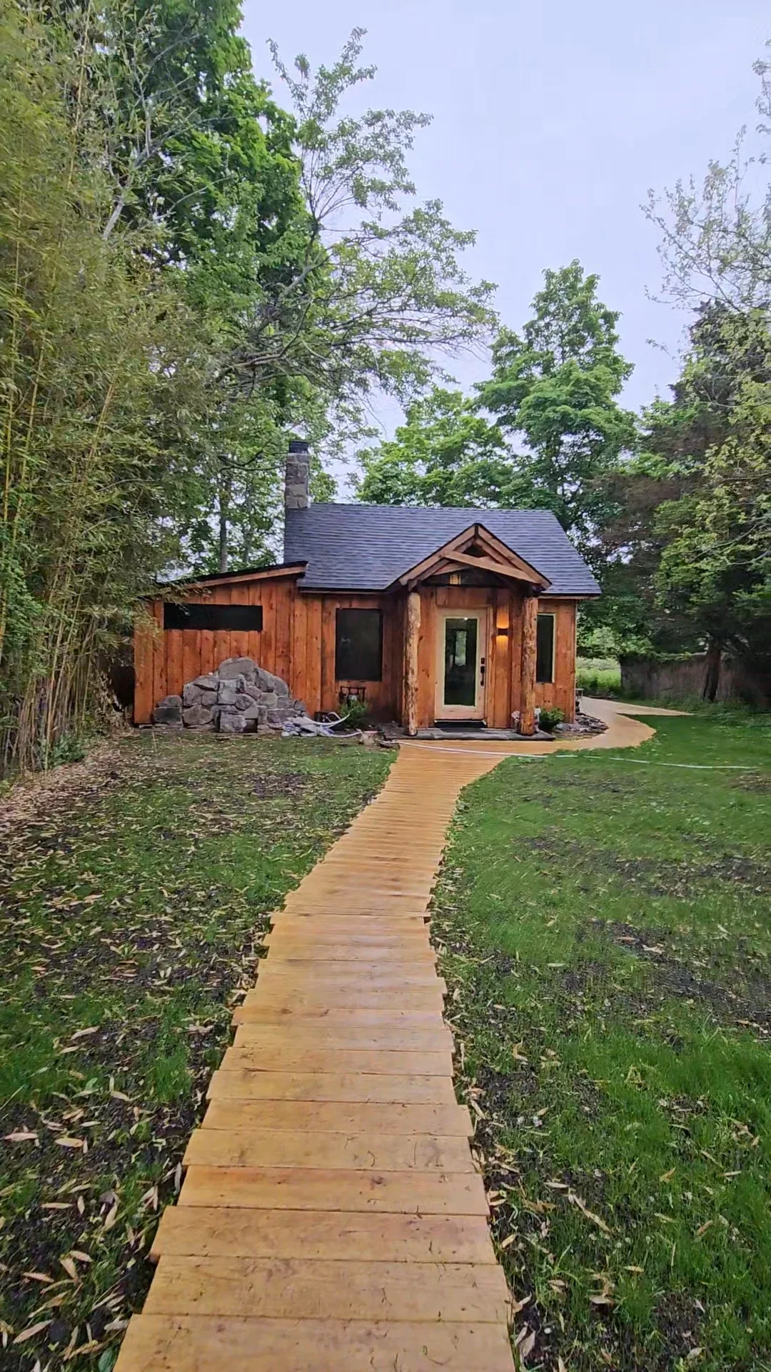 Cedar wood cabin with dark roof, brick chimney, and wooden pathway through landscaped yard
