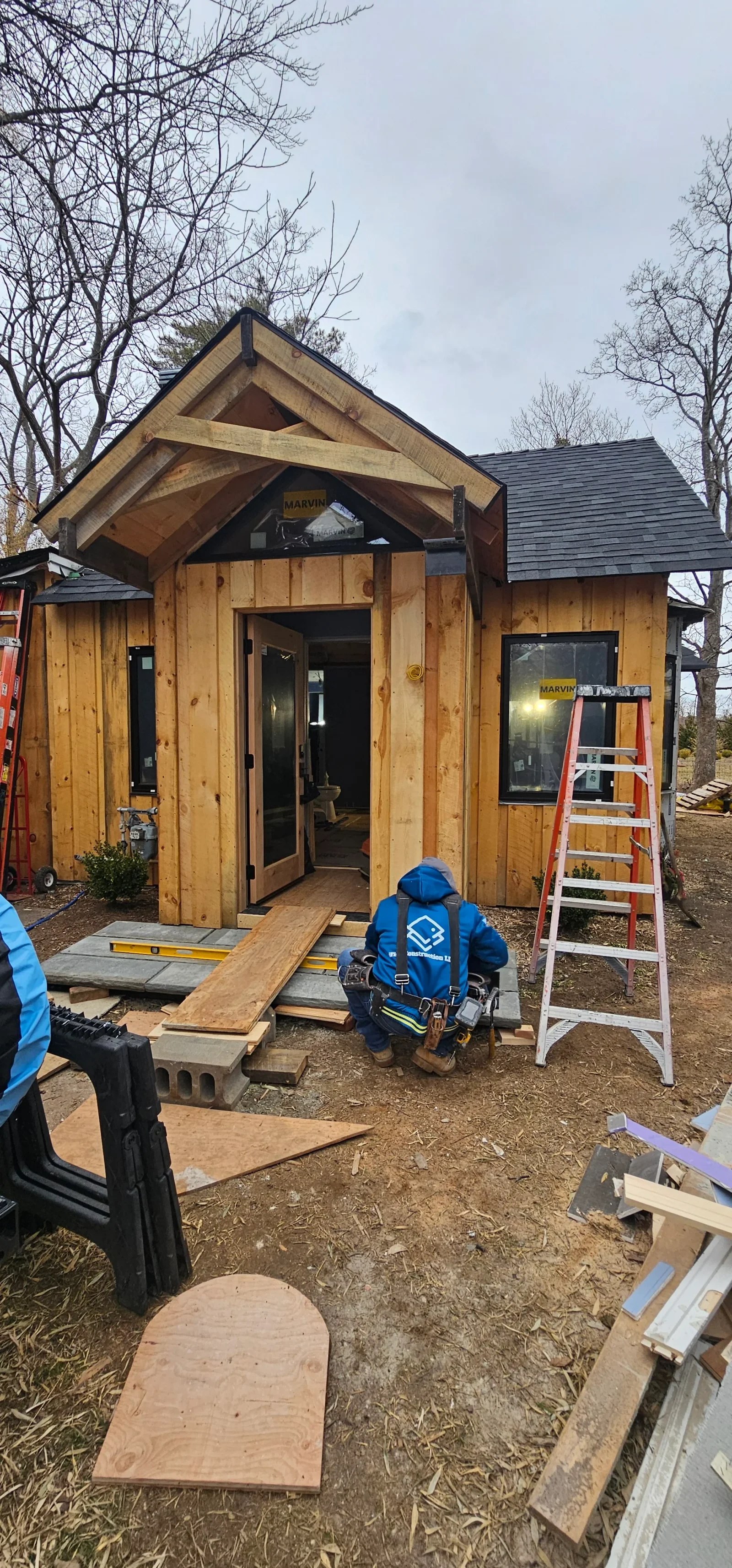 Wooden tiny home with peaked roof, natural wood siding, black trim, and wooden deck entry under construction