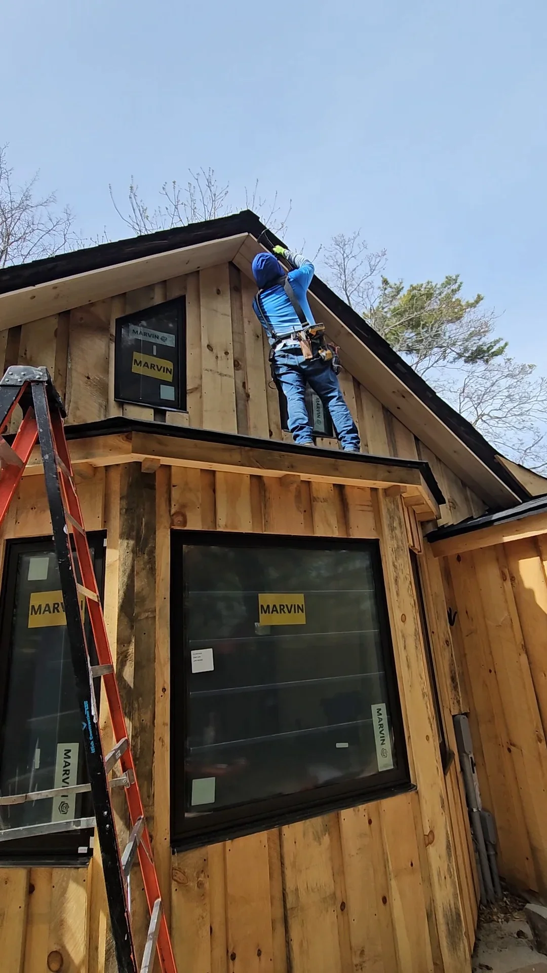 Worker installing black-framed window in wood gable end with safety harness